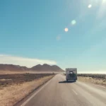 Camping-car vu de dos sur une longue route droite, entouré de plaines désertiques et de montagnes, sous un ciel bleu et un soleil intense.