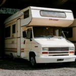Camping-car blanc « Eleanor » sous un toit métallique sur un sol de gravier.