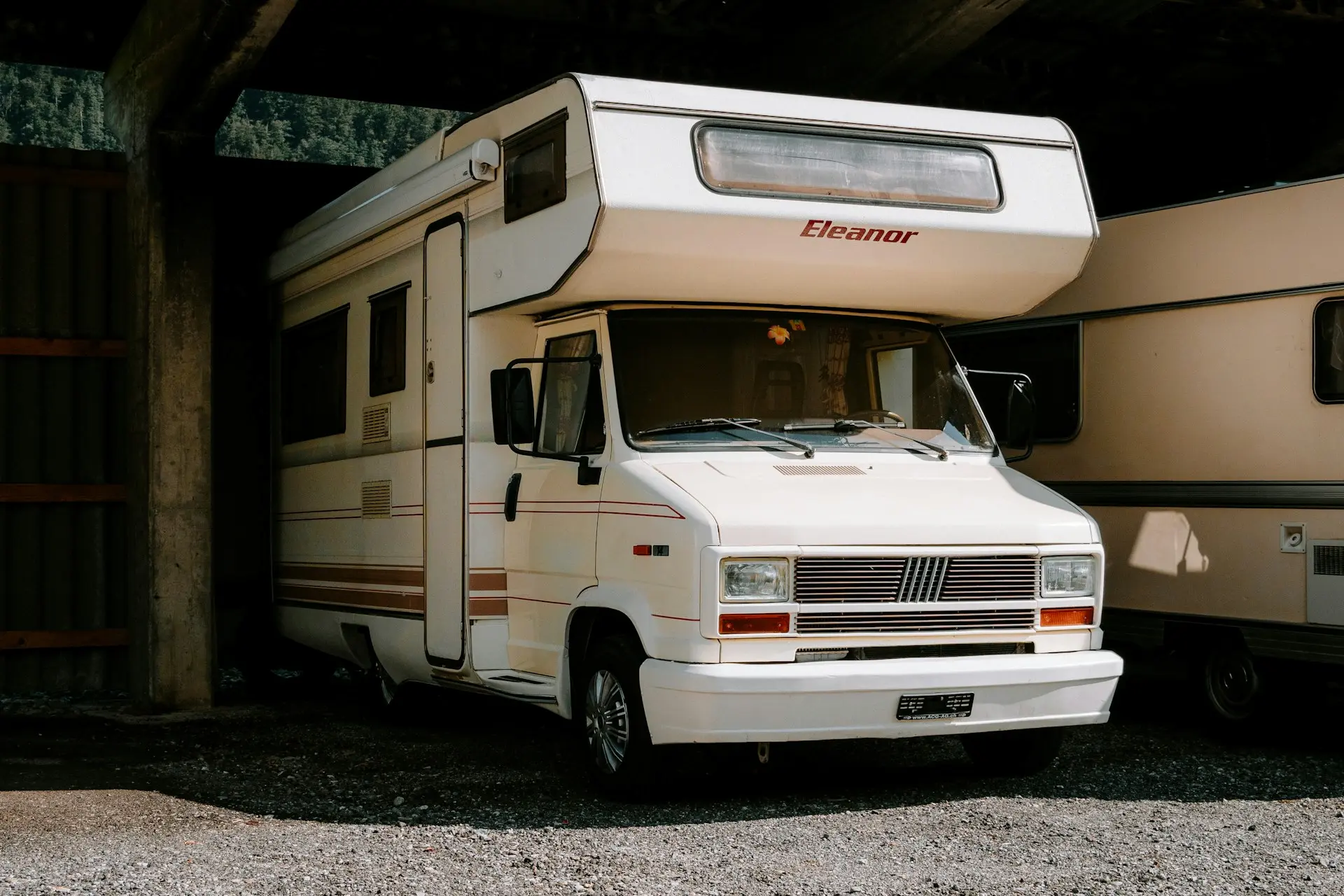 Camping-car blanc « Eleanor » sous un toit métallique sur un sol de gravier.